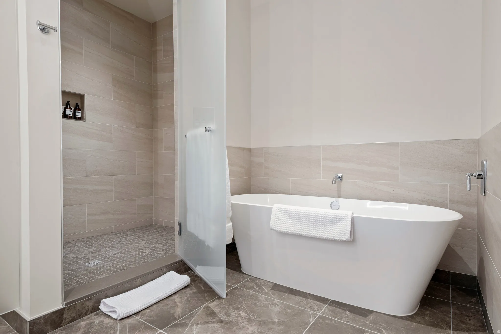 Freestanding soaking tub alongside a walk-in shower with glass paneling, tile flooring, and a tiled shower niche