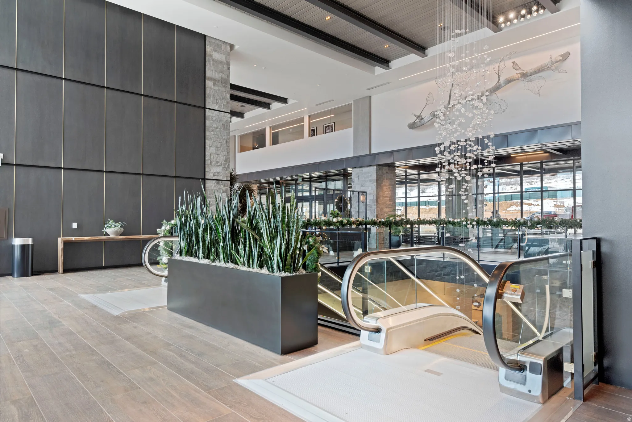 Multi-level lobby featuring wood-finish flooring, a contemporary chandelier, a stone-clad column, and an escalator with glass railings