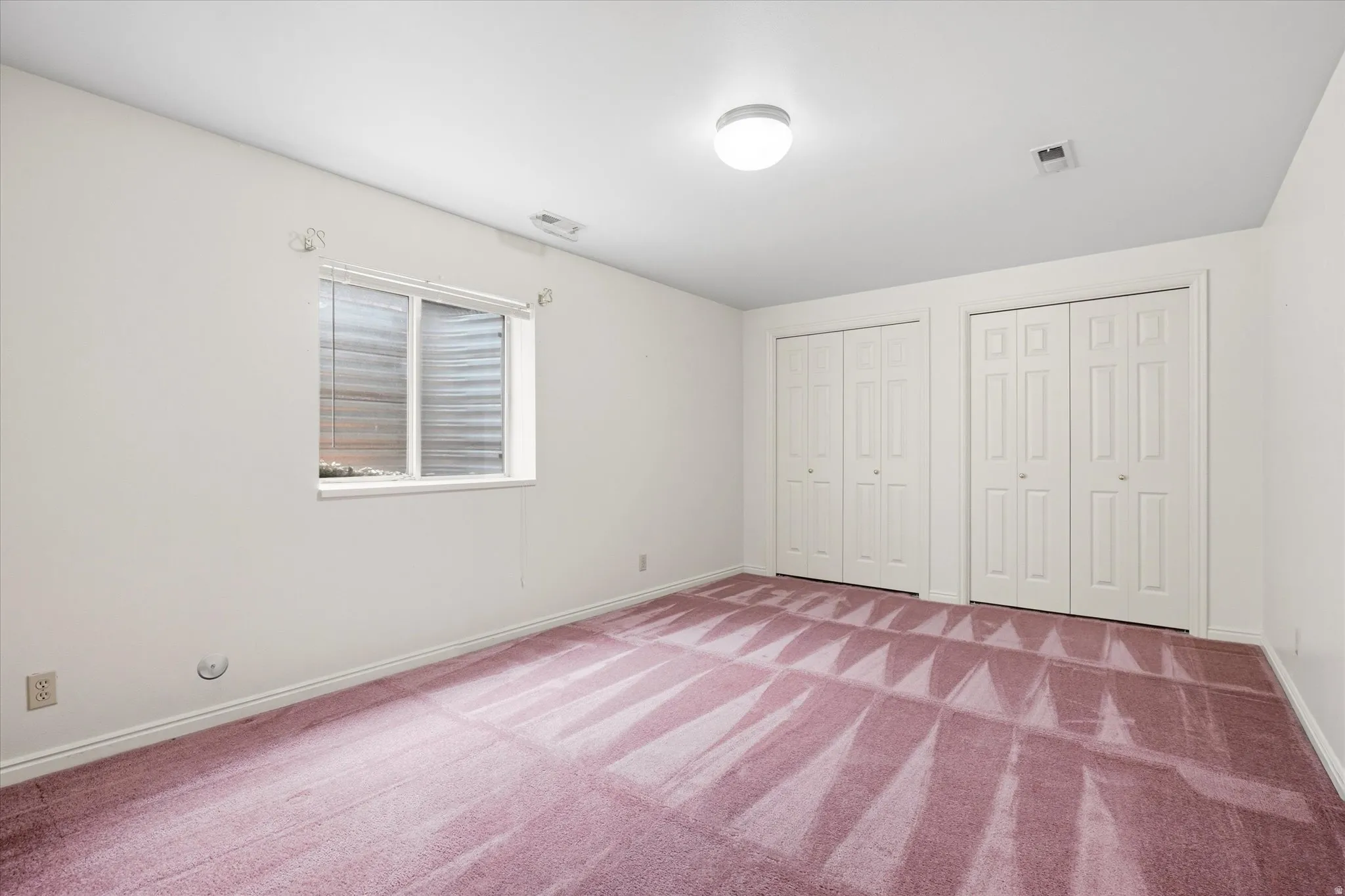 Spacious room featuring two bi-fold door closets, recessed window with blinds, overhead lighting, and carpeted flooring