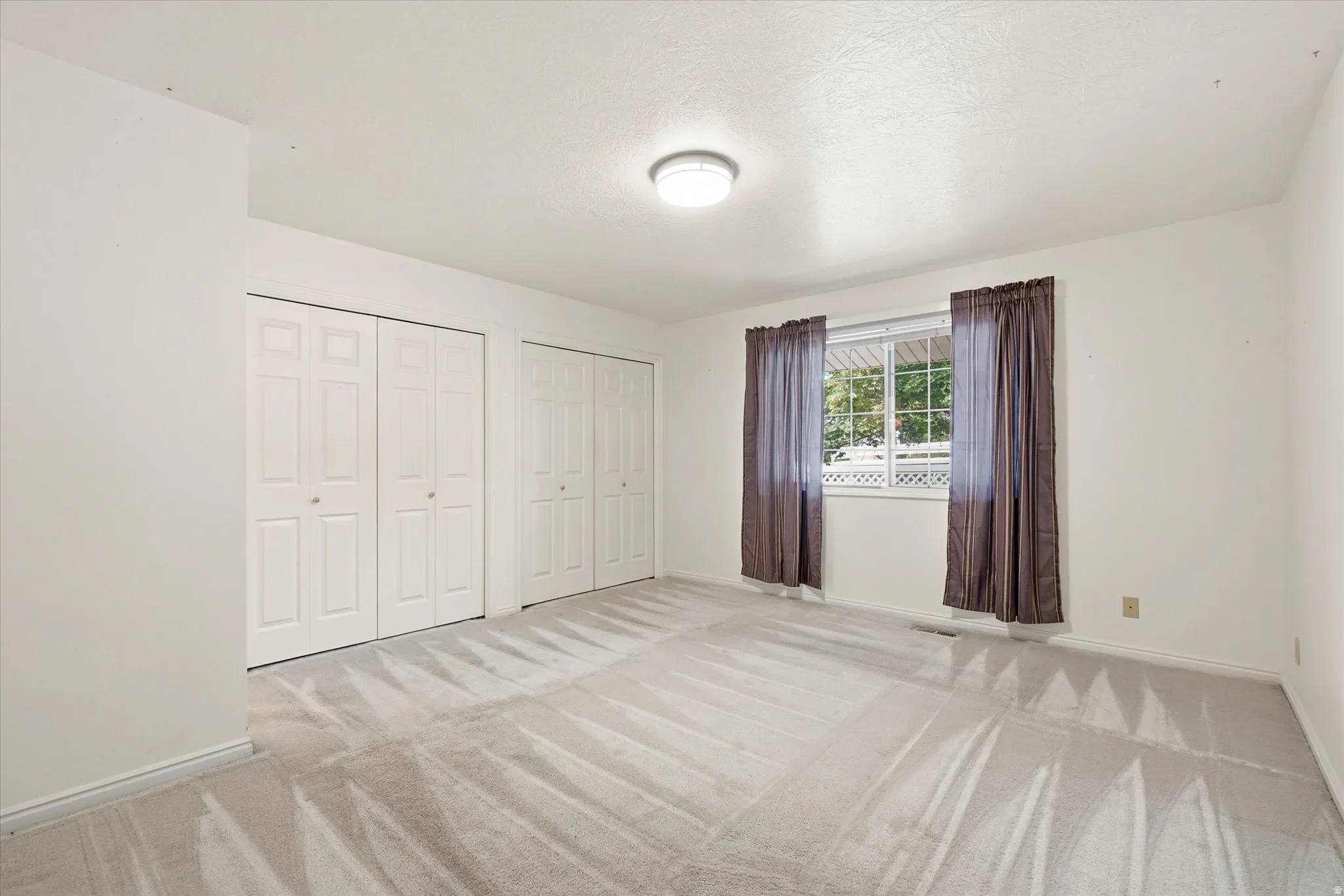 Spacious room featuring light-colored carpeting, multiple bifold closet doors, a window with exterior visibility, and a ceiling-mounted light fixture