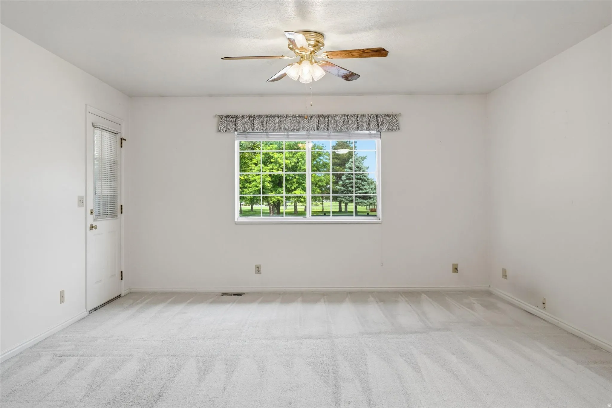 Spacious room featuring light-colored carpeting, a window with exterior views, a ceiling fan with integrated lighting, and a white paneled door