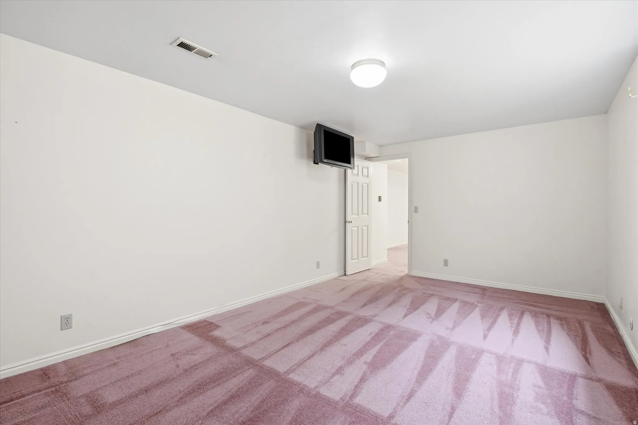 Spacious room featuring light-toned walls, a flush-mount ceiling light fixture, and a wall-mounted built-in television