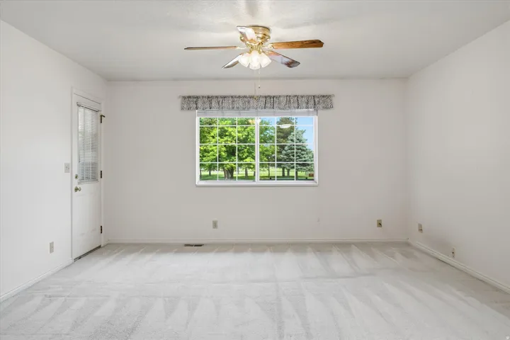 Spacious room featuring light-colored carpeting, a window with exterior views, a ceiling fan with integrated lighting, and a white paneled door