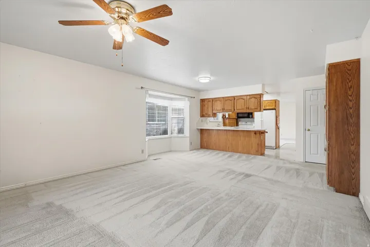 Spacious carpeted room featuring a bay window, ceiling fan, and an integrated kitchen area with wood-finish cabinetry, white countertops, and a breakfast bar