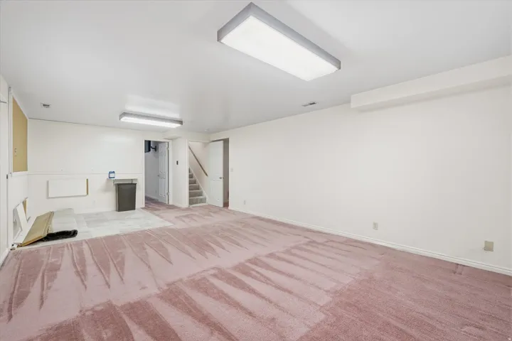 Spacious room featuring carpeting, white walls, overhead fluorescent lighting, and visible staircase access