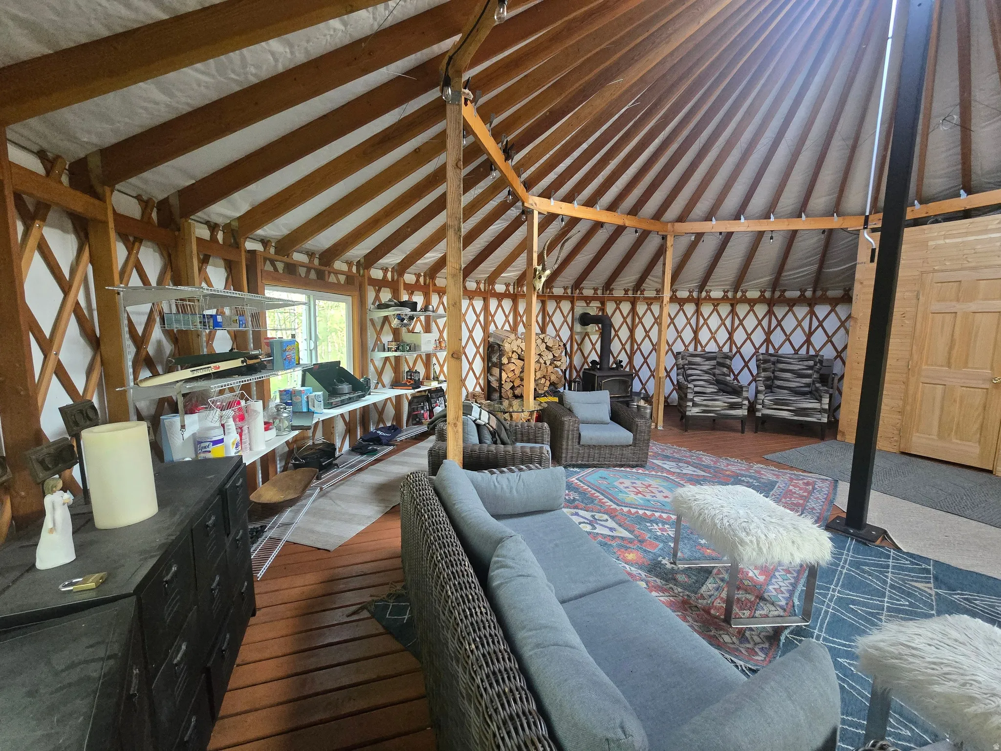 Expansive circular interior featuring exposed wooden rafters, lattice wall detailing, wood-finish flooring, a freestanding wood-burning stove, and a natural wood paneled door