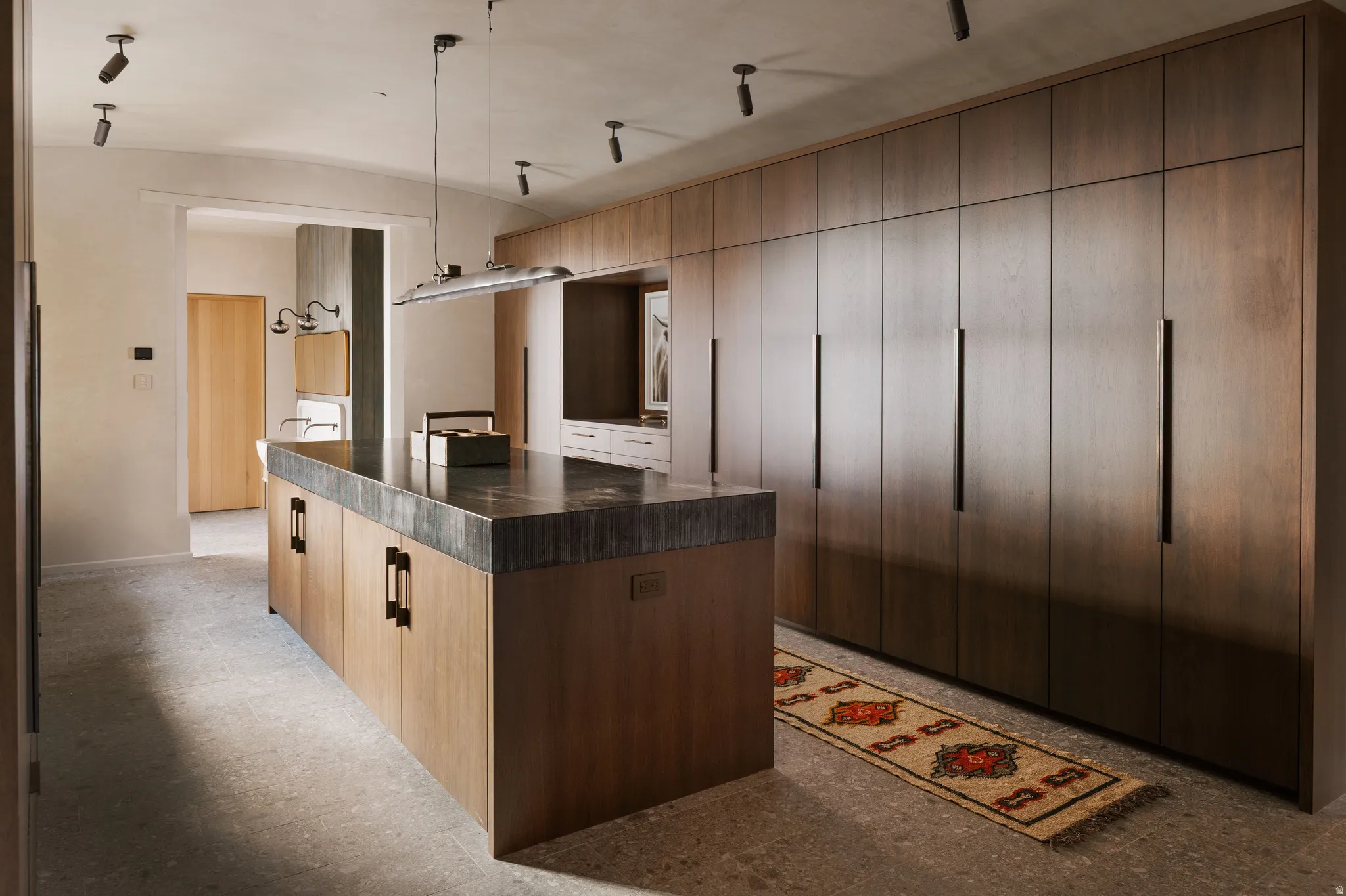 Spacious dressing room featuring an extensive wall of custom-built wood cabinetry with long black pulls, a large central island with a dark stone countertop, and recessed ceiling lighting