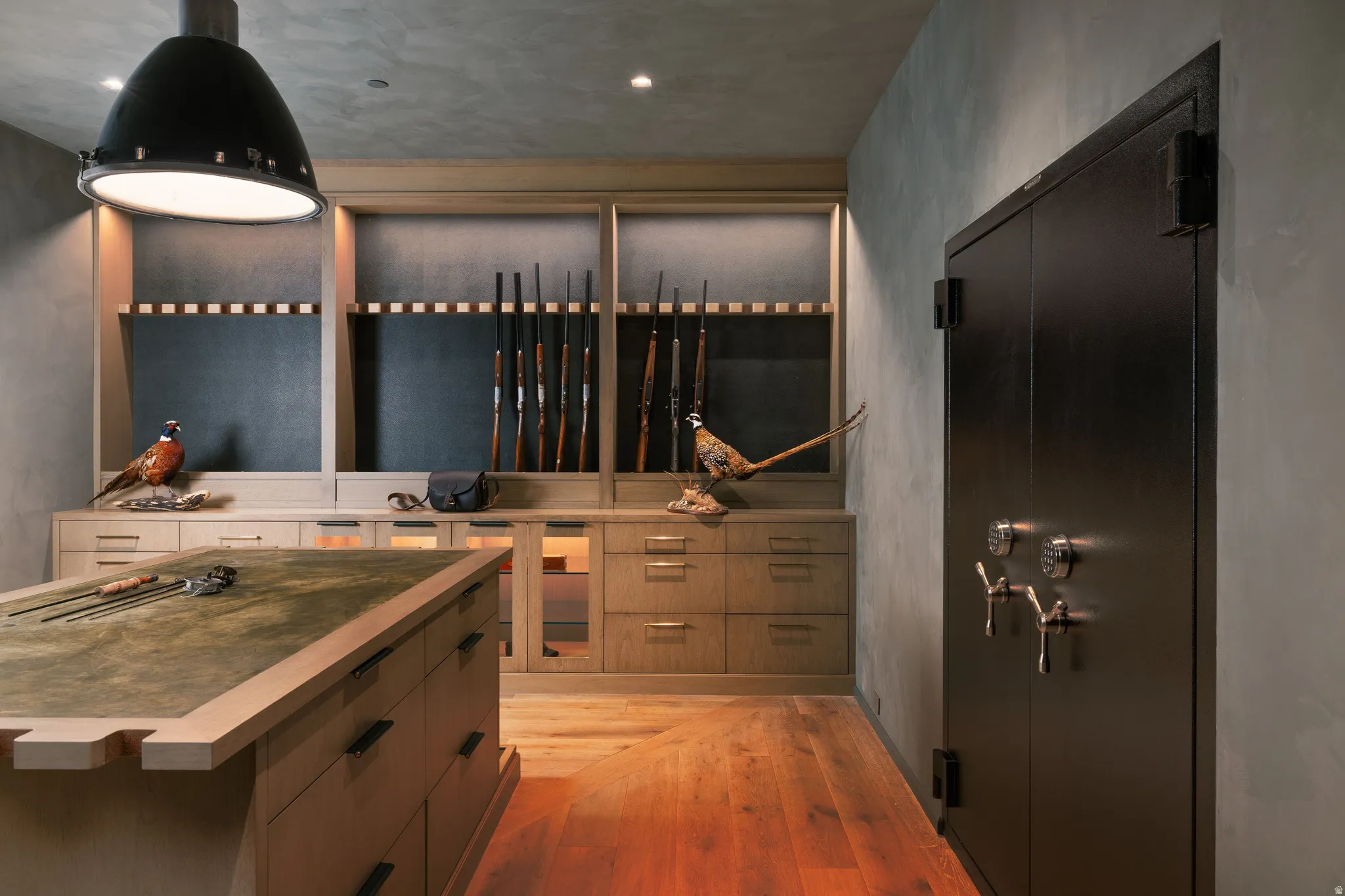 Custom-built storage room featuring a substantial black vault door, extensive light wood cabinetry with display shelving, a central work island with a green felt surface, and warm wood-finish flooring