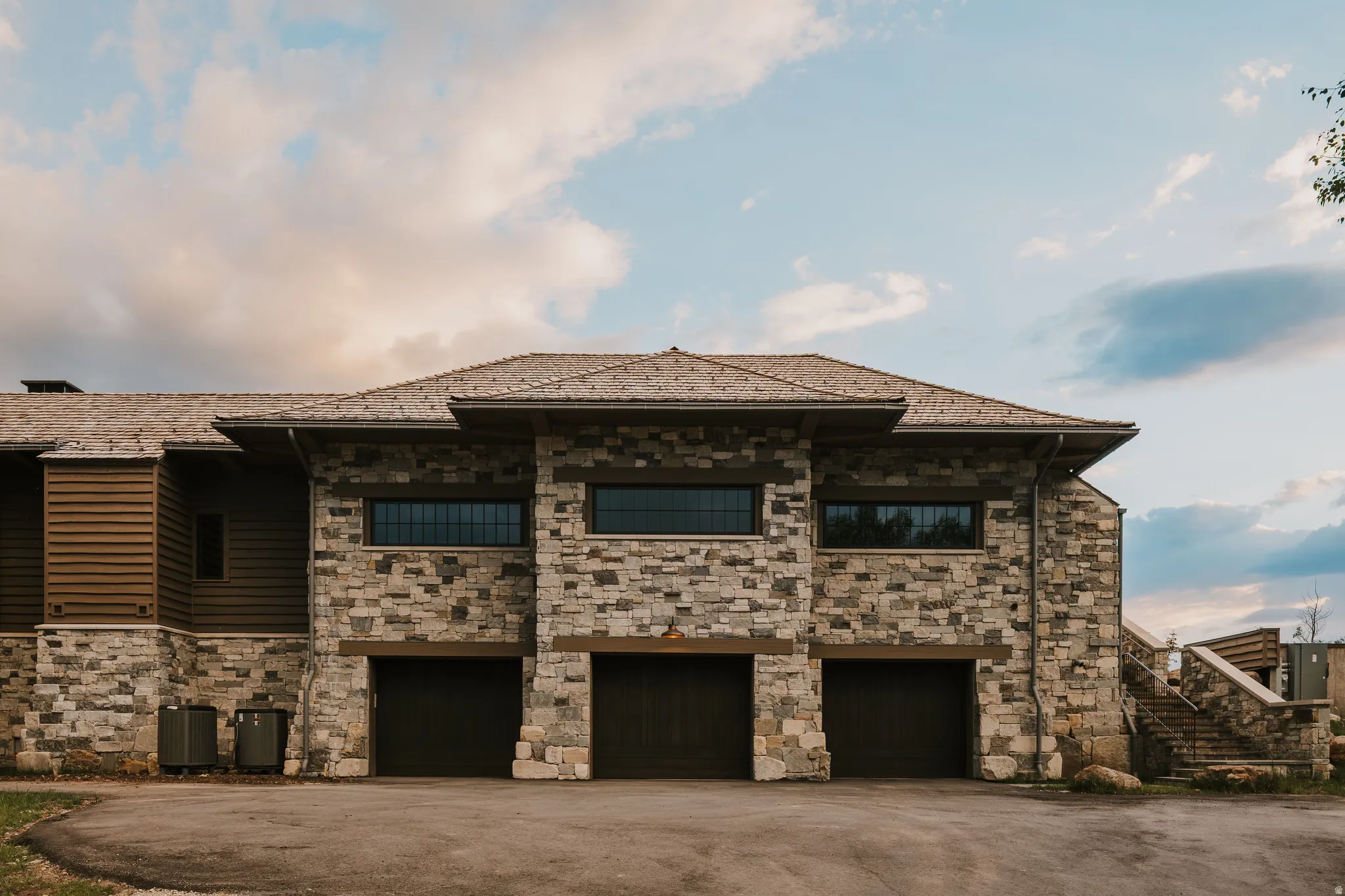 Stone and wood-sided exterior featuring three garage bays, a shingle roof, and a stone staircase