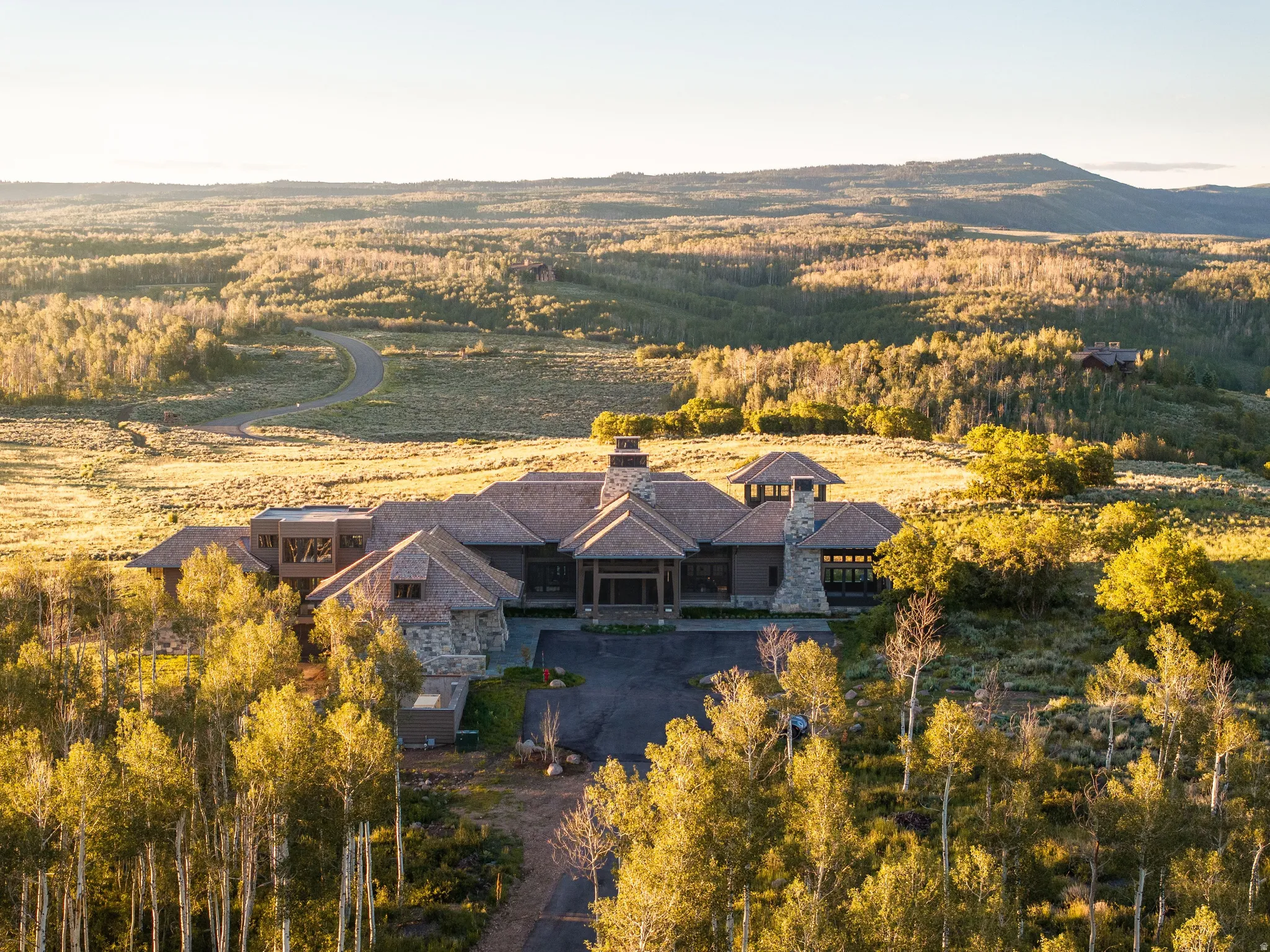 Expansive property featuring a stone and wood exterior, multiple gabled roofs, a prominent stone chimney, and an asphalt driveway, surrounded by mature trees and natural landscaping