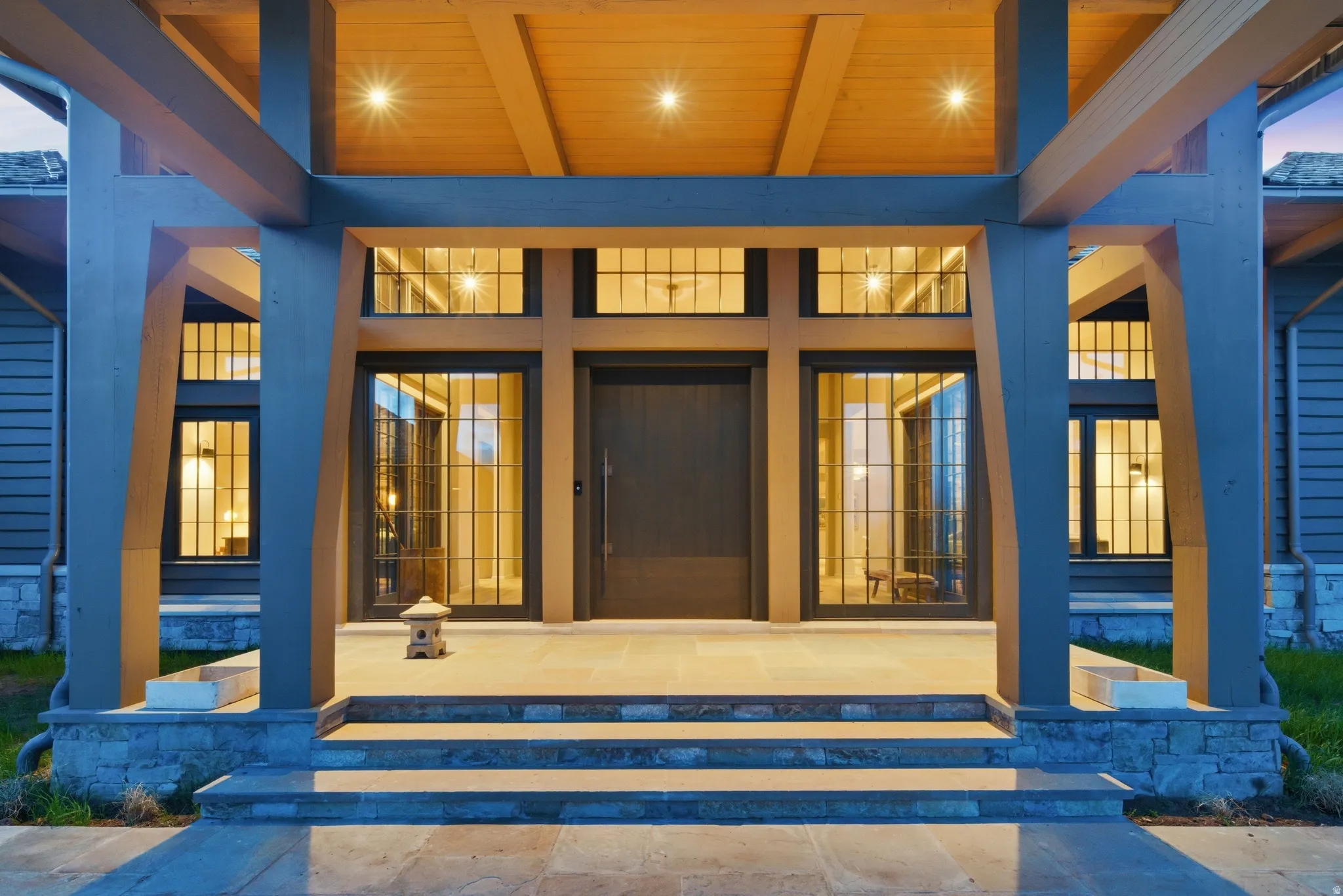 Expansive covered entryway featuring robust timber framing, a natural wood ceiling with recessed lighting, and a dark-paneled entry door