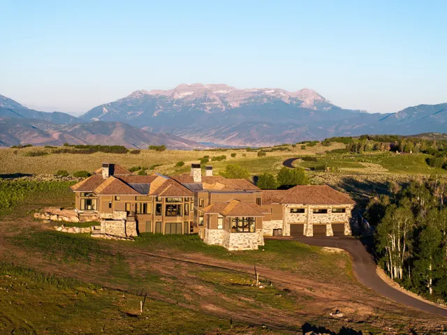Expansive estate featuring a stone and stucco exterior, multiple large windows, a stone-clad multi-car garage, and a winding paved driveway