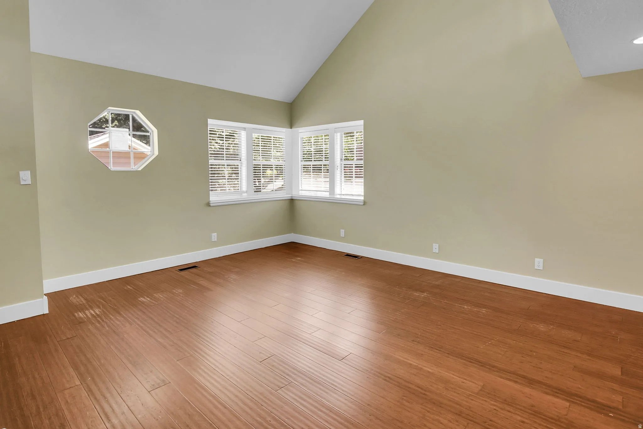 Spacious room featuring vaulted ceilings, wood-finish flooring, white baseboards, and a corner window arrangement with horizontal blinds