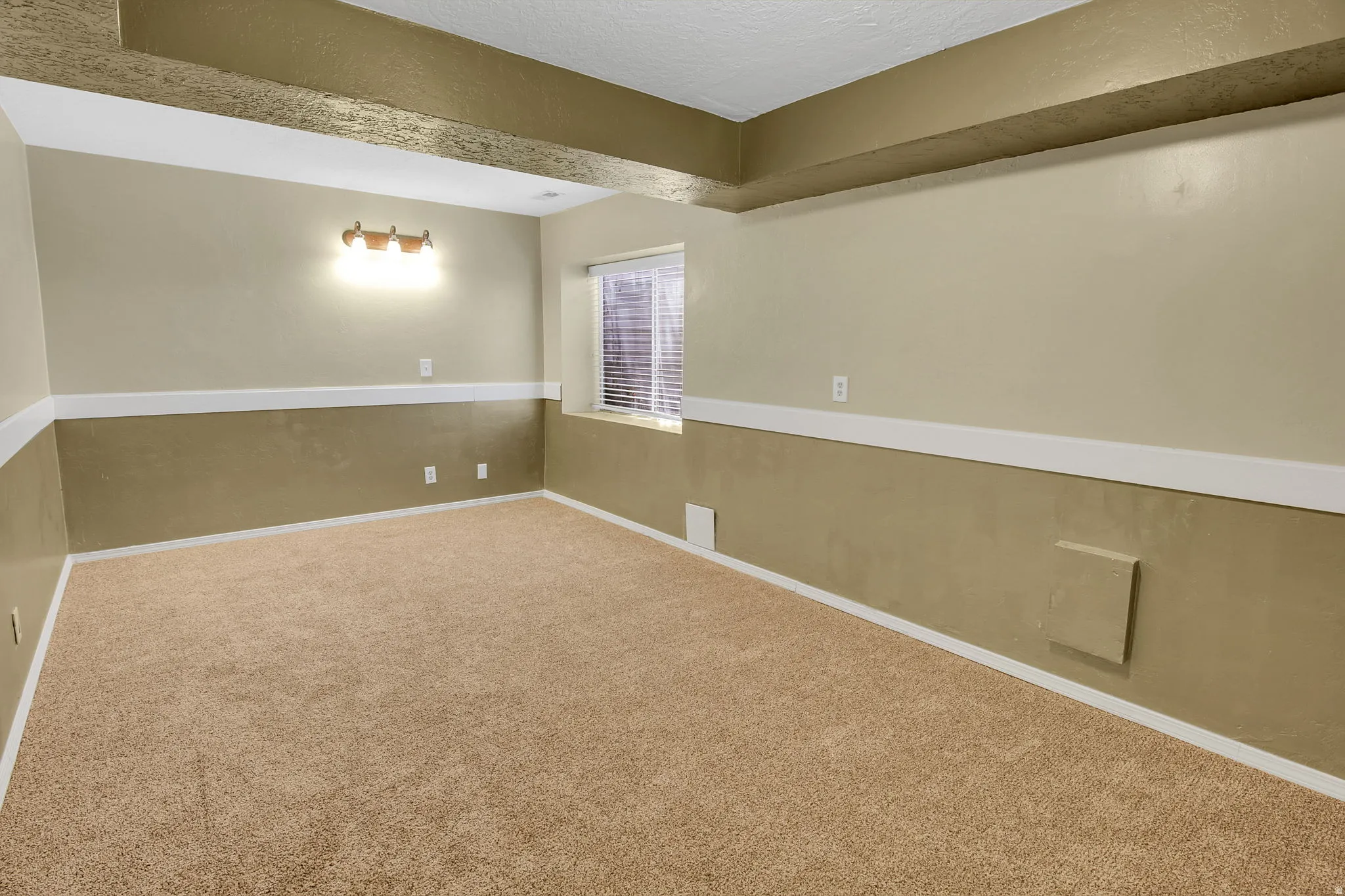 Carpeted room with a drop ceiling featuring exposed beams, two-tone painted walls with white trim, a single window with horizontal blinds, and a wall-mounted light fixture