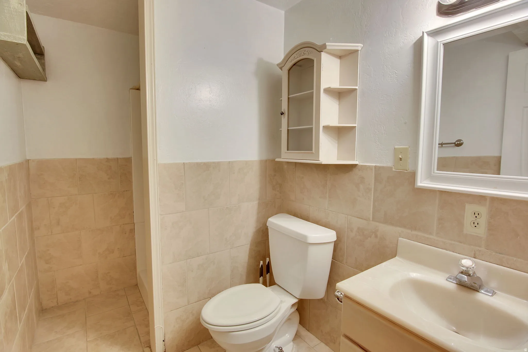 Bathroom featuring floor-to-ceiling tiled shower, light-toned wall tiling, white vanity with integrated sink, framed wall mirror, and wall-mounted storage cabinet
