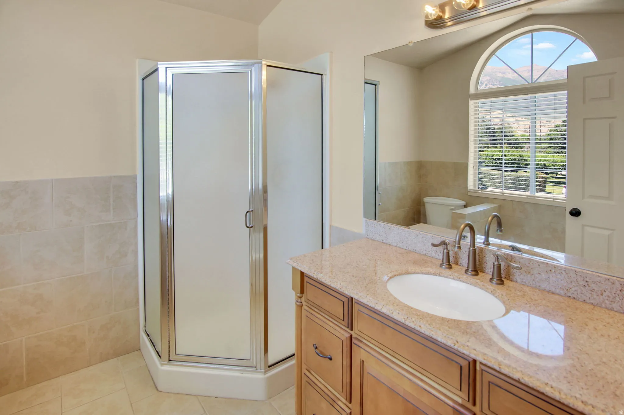 Neutral-toned bathroom featuring a corner shower with frosted glass, a single vanity with a light granite countertop, an undermount sink, and a large arched window with horizontal blinds