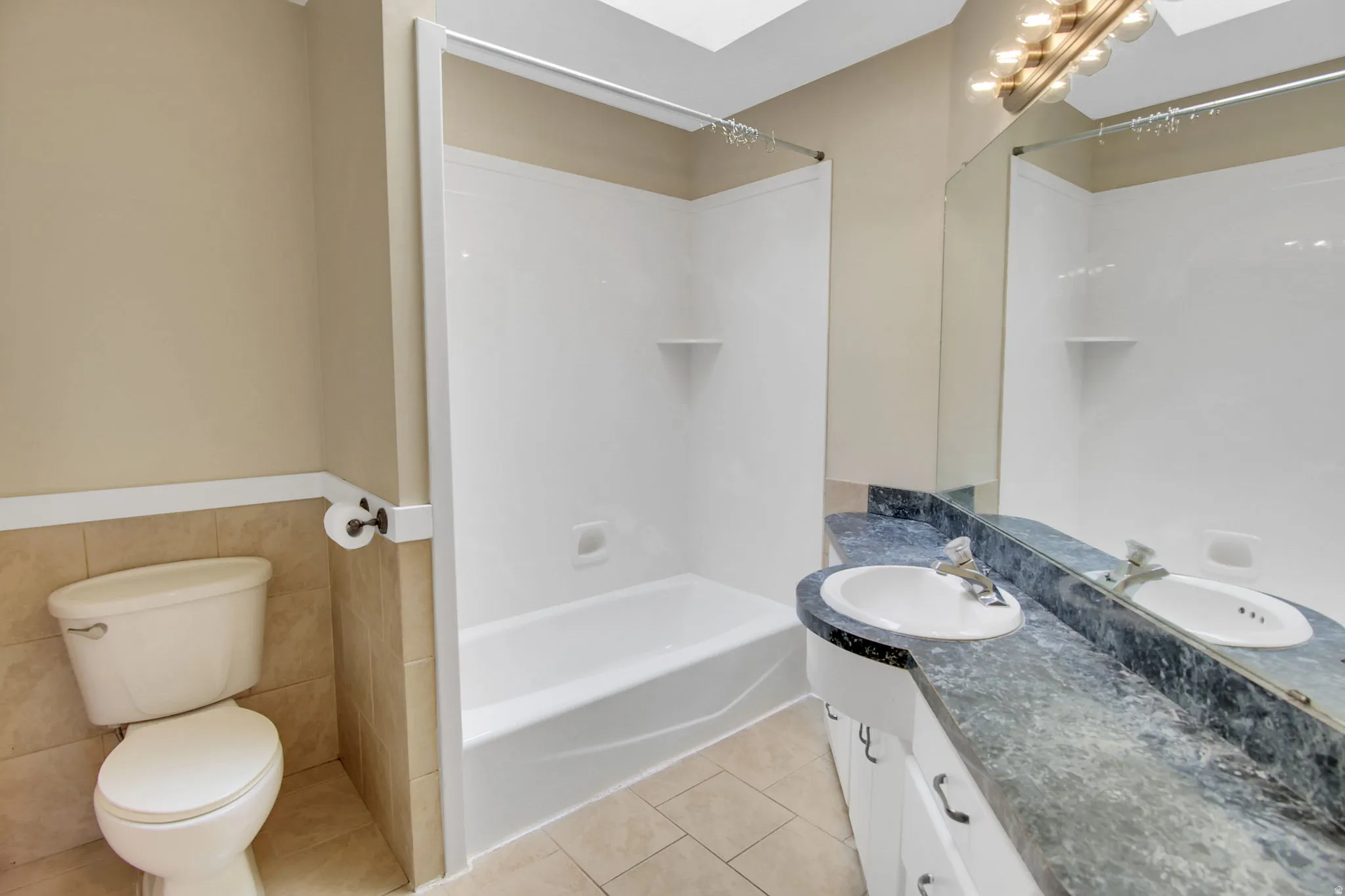 Bathroom featuring a combination tub and shower with white surround, a toilet, a vanity with an integrated sink, a dark patterned countertop, and a large mirror