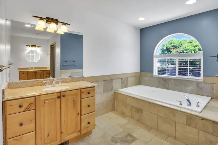Spacious bathroom featuring a built-in soaking tub with tile surround, an arched window, and a wood vanity with a light-toned countertop