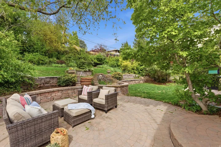 Expansive backyard patio featuring circular paver stones, multiple stone retaining walls, and surrounding mature trees and shrubbery