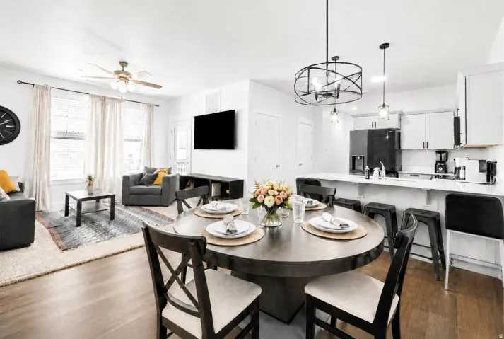 Open-concept living space with wood-finish flooring, a contemporary kitchen featuring white cabinetry and a breakfast bar, and a dining area illuminated by a modern chandelier