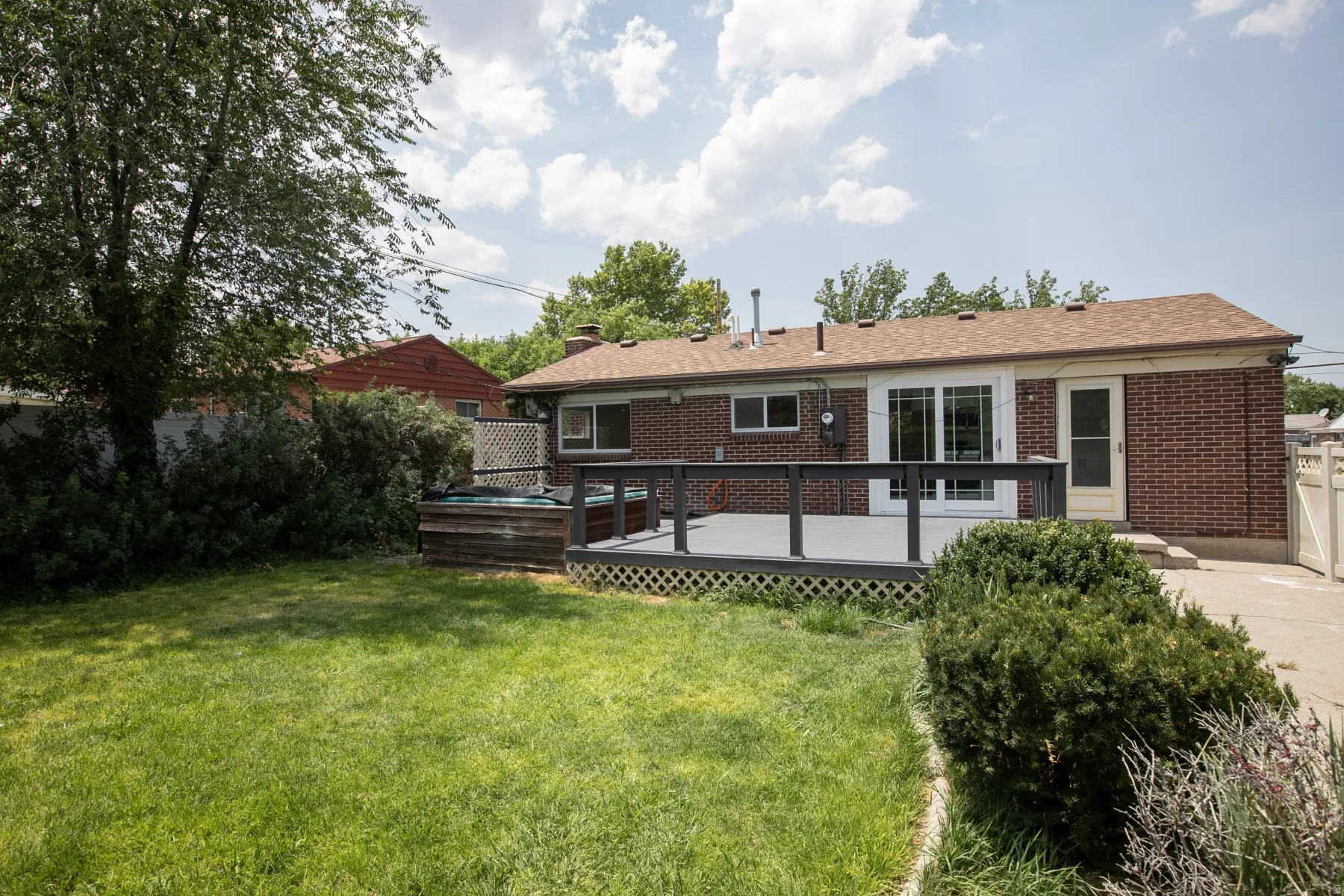 Expansive backyard featuring a manicured lawn, a spacious deck with dark railings, a built-in outdoor spa, a red brick exterior, and a white privacy fence