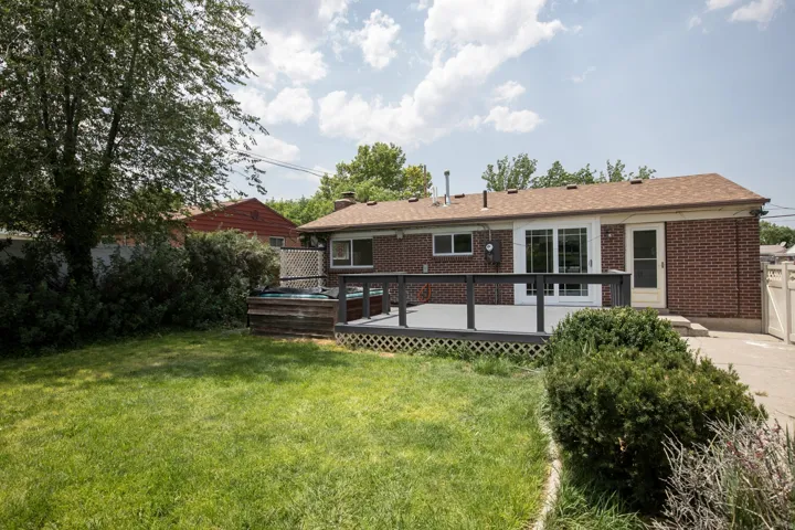 Expansive backyard featuring a manicured lawn, a spacious deck with dark railings, a built-in outdoor spa, a red brick exterior, and a white privacy fence