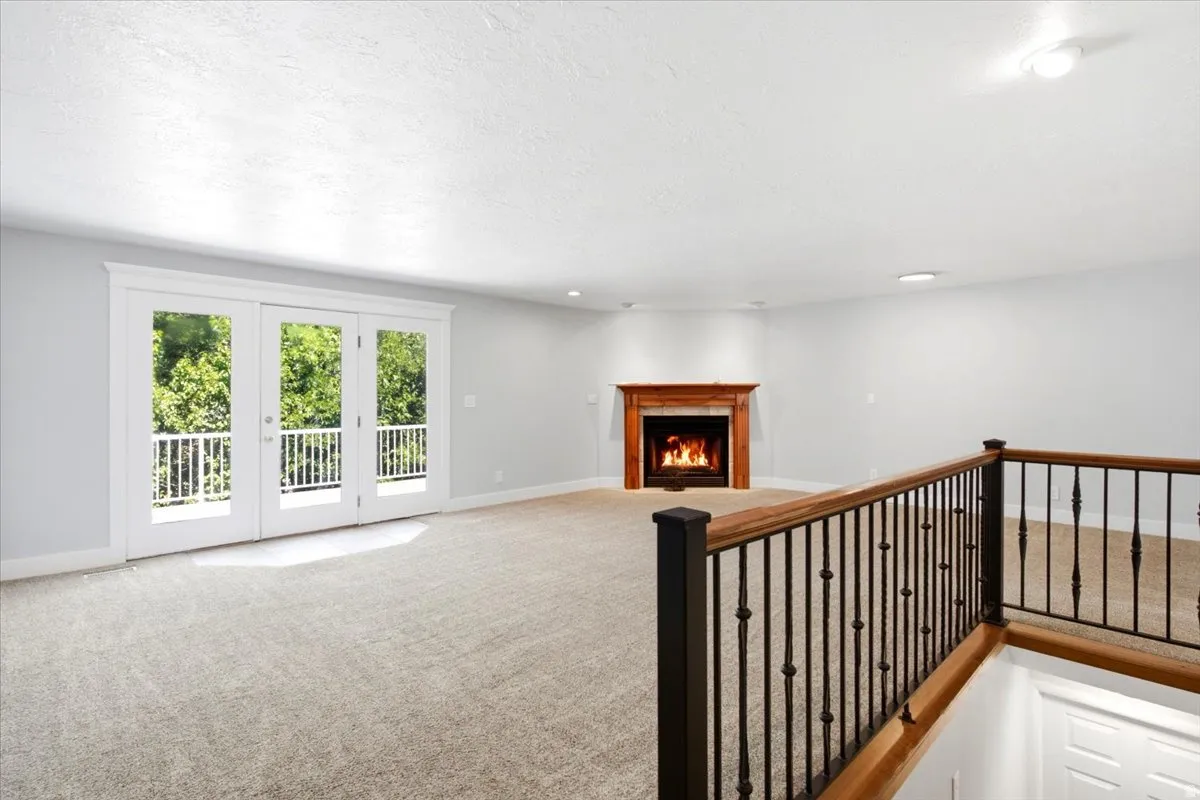 Spacious room featuring light-colored carpeting, a corner fireplace with a wood mantel, and triple French doors opening to a balcony with white railings