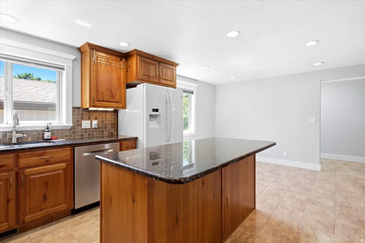 Kitchen featuring a central island with a dark countertop, wood cabinetry, stainless steel dishwasher, tile flooring, and recessed lighting