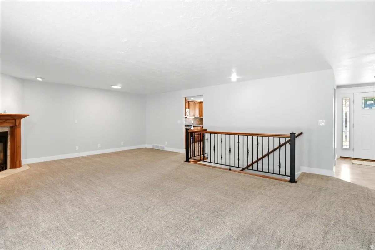 Spacious carpeted living area featuring a corner fireplace with a wooden mantel, recessed lighting, and a dark iron and wood-finish railing