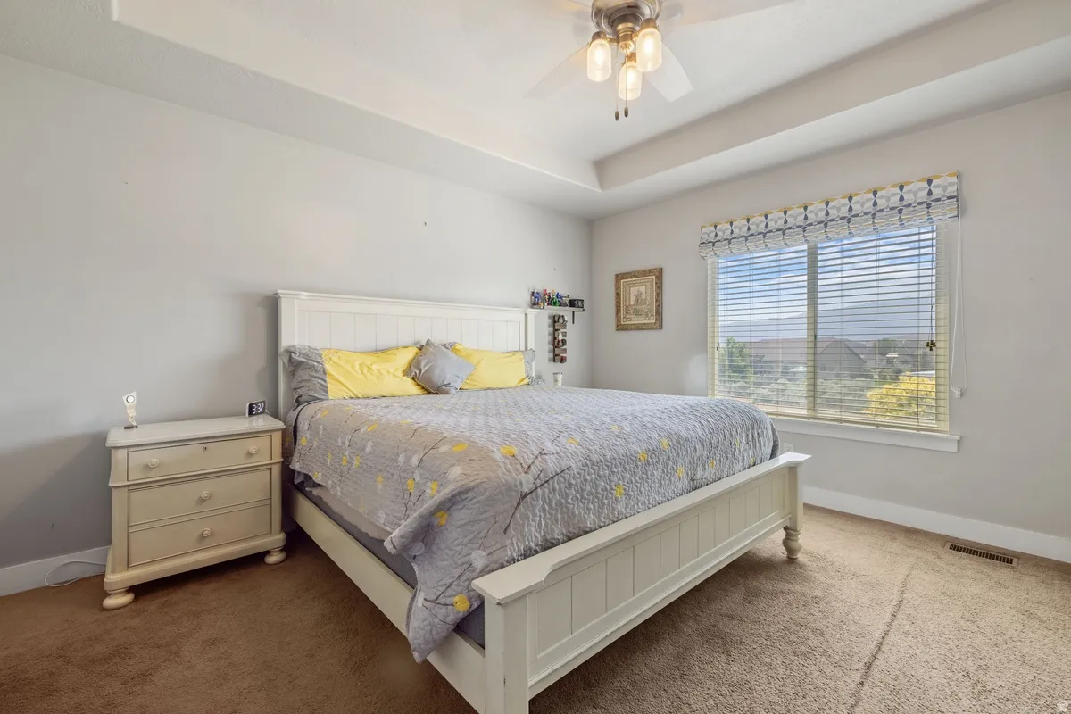 Spacious bedroom featuring a tray ceiling, ceiling fan, large window with horizontal blinds, and carpeting