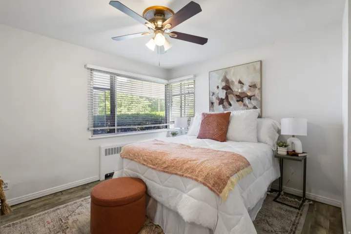 Main bedroom with wood-finish flooring, a corner window with horizontal blinds, a white baseboard heater, and a ceiling fan with integrated lighting