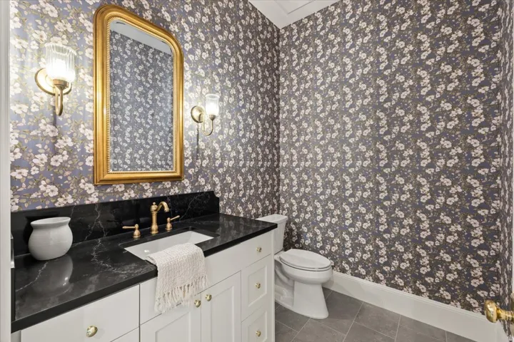 Powder room featuring floral wallpaper, a white vanity with gold hardware, a black countertop, gold-finish wall sconces, and a gold-framed mirror