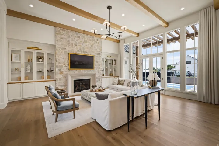 Spacious living area featuring a stone-clad fireplace with a mantel, built-in cabinetry, exposed ceiling beams, and wood-finish flooring