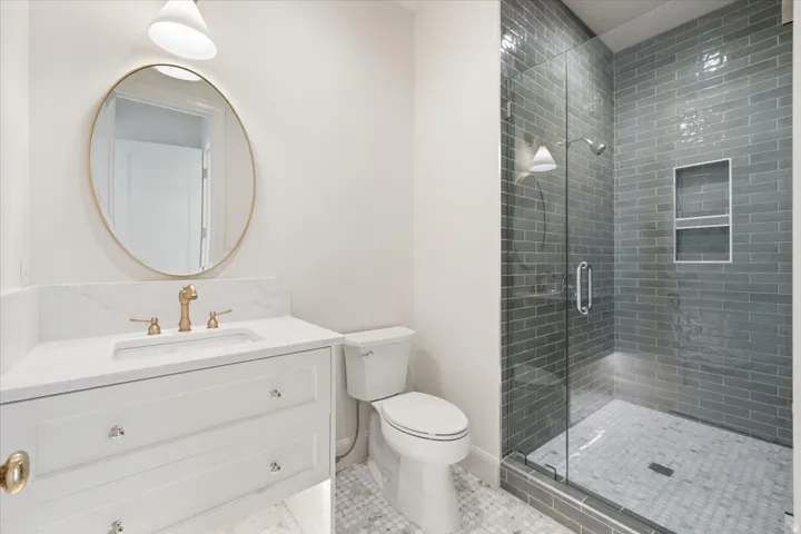Bathroom featuring a walk-in shower with subway tile surround and built-in niches, glass shower enclosure, vanity with stone countertop, undermount sink, and brass-finish faucet, and a round frameless mirror