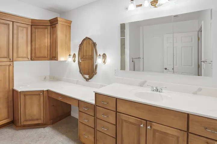 Spacious bathroom vanity featuring light wood cabinetry, white countertops, an integrated sink with a chrome faucet, a large mirrored medicine cabinet, and an additional ornate oval mirror with sconce lighting