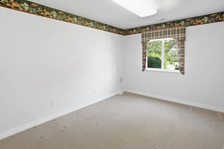 Room featuring a single window with a plaid valance, light beige carpeting, white walls, white baseboards, and a decorative wallpaper border