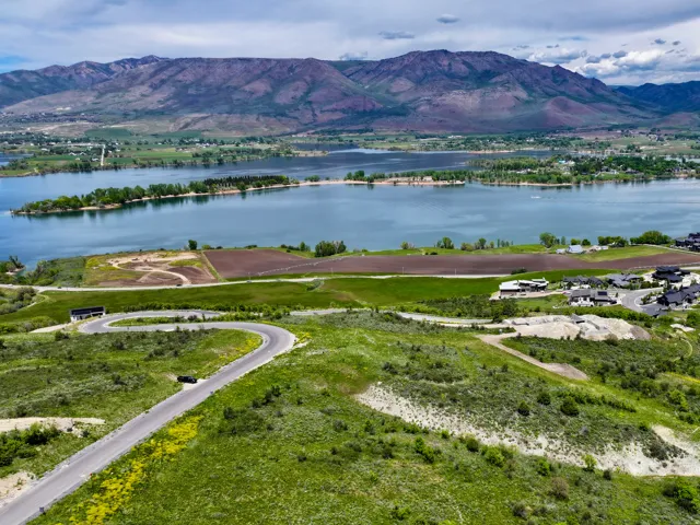 Expansive green landscape with a winding paved road, featuring a prominent lake backdrop with distant mountains, and a community of modern homes