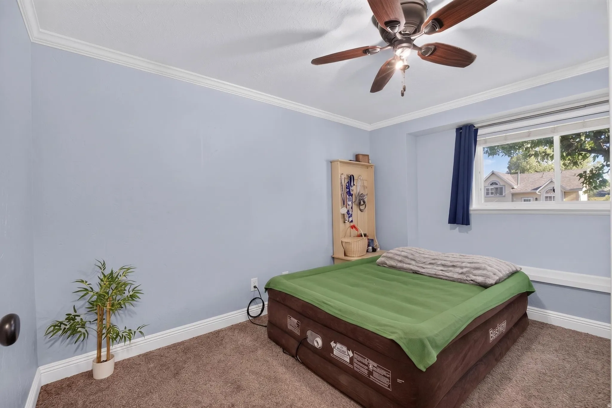 Bedroom featuring a ceiling fan with integrated lighting, a window with a white frame, light blue walls, white crown molding, and brown carpet flooring
