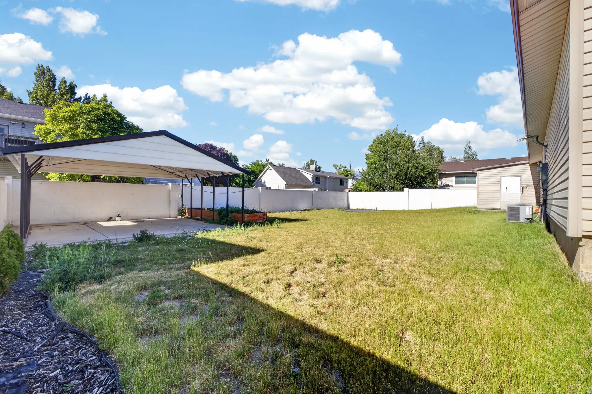 Expansive rear yard featuring a covered carport structure, concrete pad, perimeter fencing, and established trees