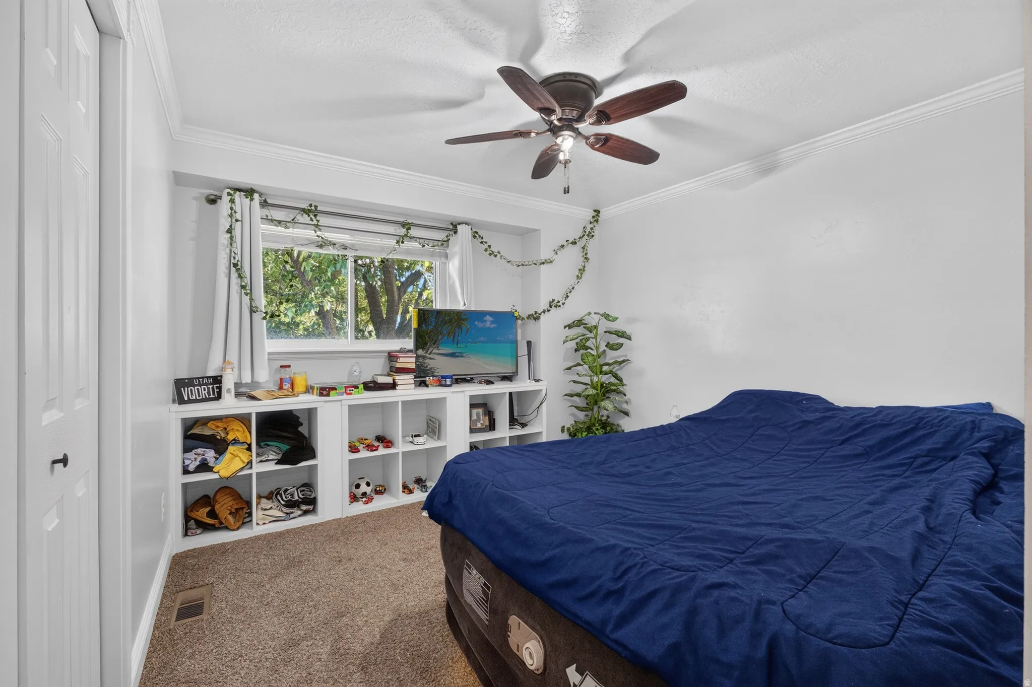 Bedroom featuring a window with exterior tree scenery, built-in shelving, crown molding, a ceiling fan, and carpet flooring