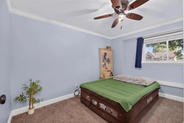 Bedroom featuring a ceiling fan with integrated lighting, a window with a white frame, light blue walls, white crown molding, and brown carpet flooring