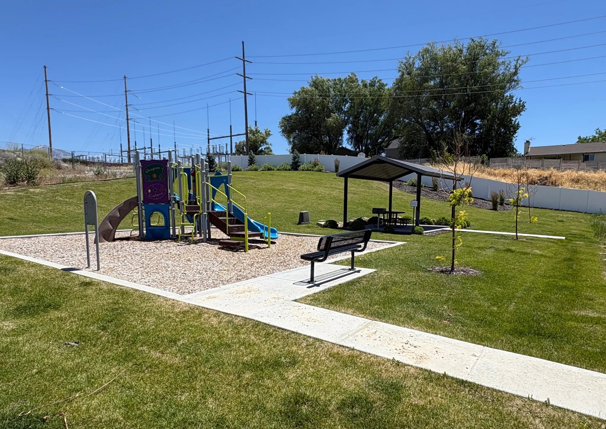 Community playground featuring a colorful play structure on a wood chip surface, shaded picnic area, and a paved walking path