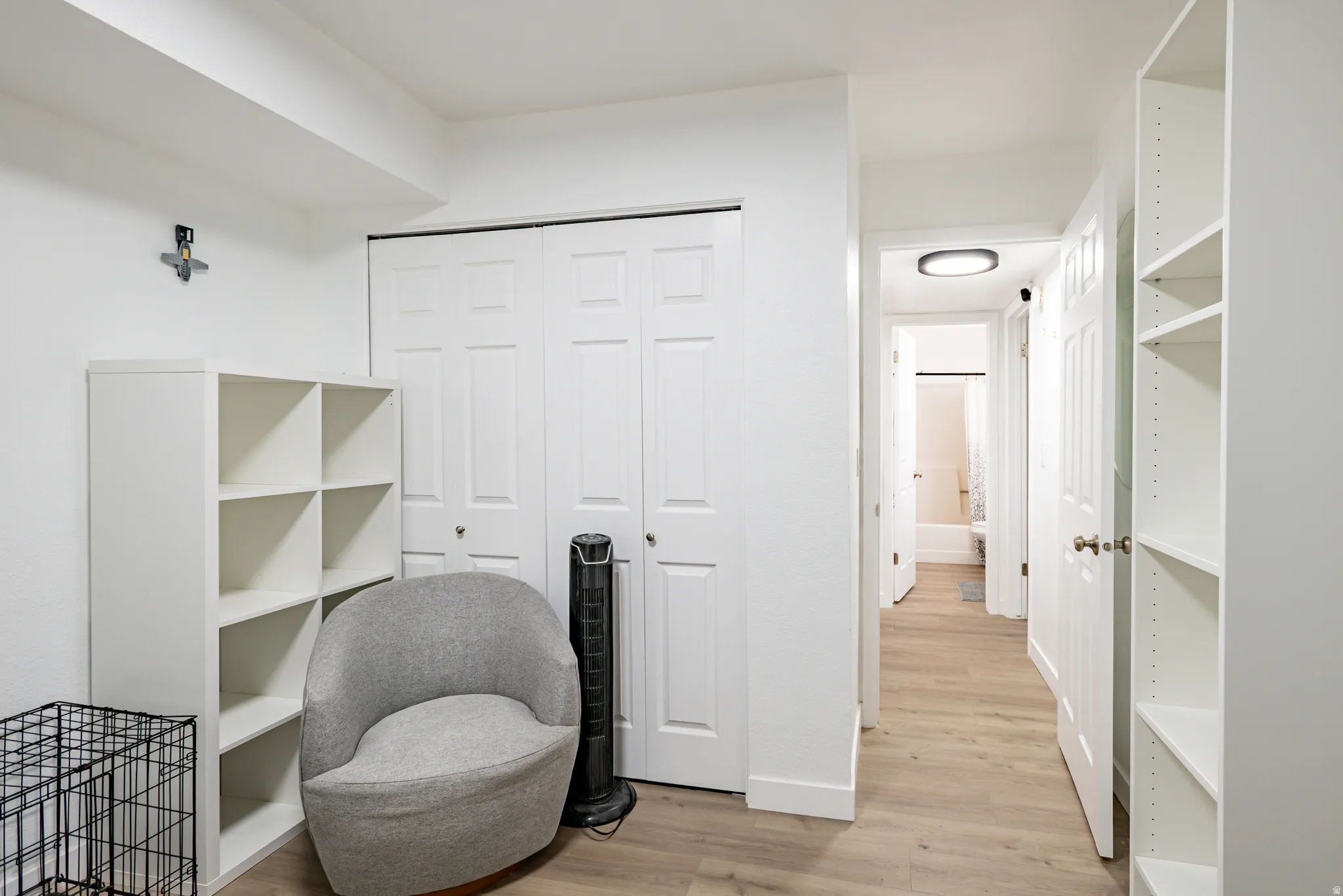 White built-in shelving, light wood-finish flooring, white paneled bifold doors, and a hallway leading to a bathroom with a white tub