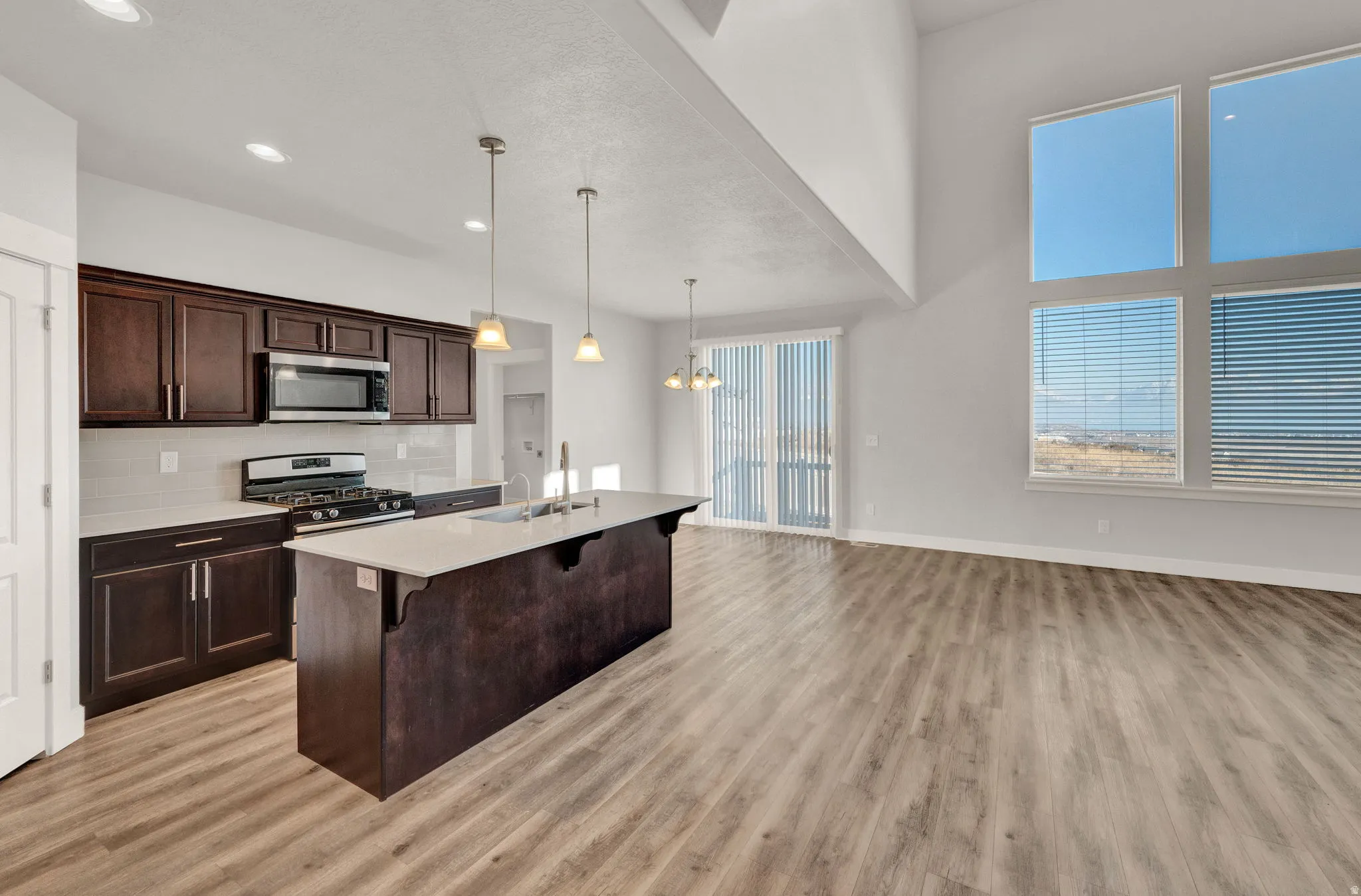 Open-concept living area featuring a kitchen island with pendant lighting, stainless steel appliances, wood-finish flooring, and large windows with blinds