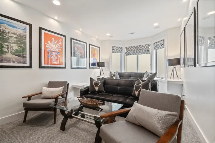 Recessed lighting illuminates this living space featuring a bay window with patterned Roman shades, carpeted flooring, and white baseboards