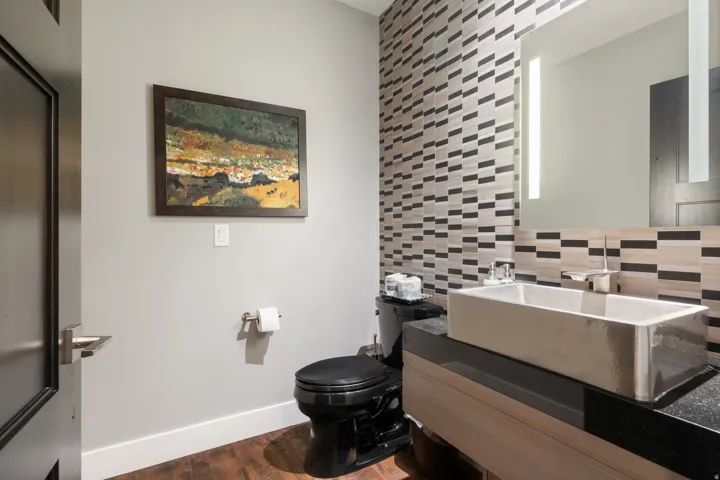 Modern bathroom featuring a rectangular vessel sink, black countertop, patterned tile accent wall, built-in light mirror, and rich wood-finish flooring
