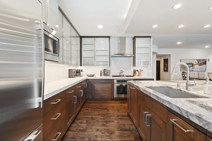 Gourmet kitchen featuring a stone island with undermount sink, stainless steel appliances, wood-finish flooring, recessed lighting, and rich wood cabinetry with glass uppers