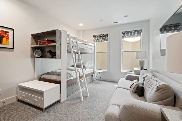 Recessed lighting illuminates this room featuring a built-in bunk bed with integrated shelving, two windows with patterned shades, and light gray wall-to-wall carpeting