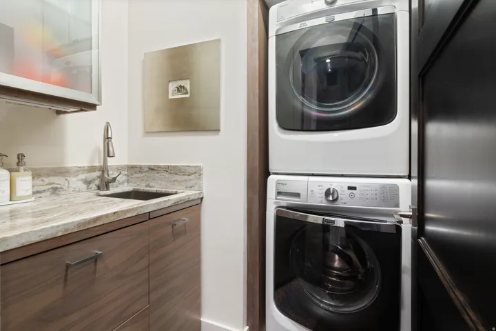 Integrated laundry area featuring stacked appliances, a stone countertop with an undermount sink, a gooseneck faucet, and wood-finish cabinetry