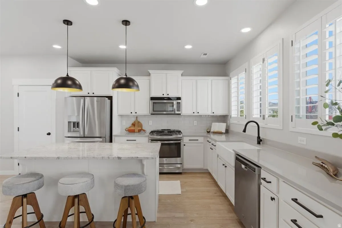 Modern kitchen featuring white shaker cabinetry, stainless steel appliances, light wood-finish flooring, a farmhouse sink with a black gooseneck faucet, and a large island with a light-colored countertop